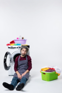 A frustrated man sitting in front of a broken washer with a laundry basket on the floorDealing with a broken washer can be overwhelming—appliance removal takes the burden off your shoulders.